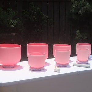 May include: A set of six pink crystal singing bowls of varying sizes, arranged on a white table cloth. The bowls are on their individual cushions and a mallet is in the foreground.