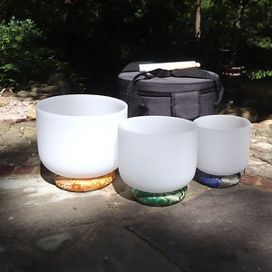 May include: Three white crystal singing bowls of different sizes, each resting on a colorful cushion. The bowls are arranged in a row on a stone patio. A black carrying case with a wooden mallet is visible in the background.