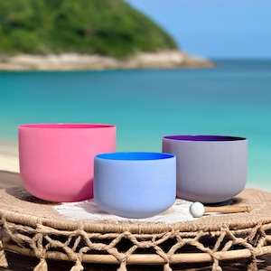 May include: Three colored crystal singing bowls in pink, blue, and gray, with a white mallet, rest on a woven mat. The bowls are on a wooden surface with a beach and ocean backdrop.