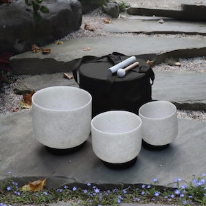 May include: Three white quartz singing bowls of varying sizes are arranged on a stone surface, with a black carrying case and two mallets in the background. The bowls have a speckled texture and sit on black bases. The scene is outdoors.