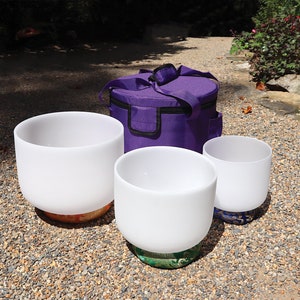 May include: Three white crystal singing bowls with a purple carrying case. The bowls are sitting on a gravel surface.