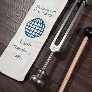 May include: A tuning fork with a wooden mallet and a white cloth bag. The bag has the words "Schumann Resonance" and "Earth Heartbeat Gaia" printed on it, along with a blue globe graphic. The tuning fork is labeled "Schumann 93.96".