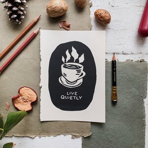 以下が含まれることがあります： 白地に、蒸気を立てたコーヒーカップと「Live Quietly」というテキストが描かれた白黒のイラストです。