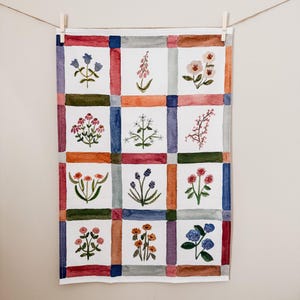 May include: A white floral tea towel with a grid pattern. Each square features a different watercolor flower design in shades of blue, pink, orange, and green. The towel is hanging from a wooden clothespin on a string.