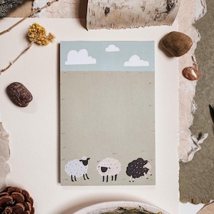 May include: A notepad with a light blue sky and white cloud design at the top, and a tan field with three sheep illustrations at the bottom. The sheep are white, off-white, and black. The notepad is surrounded by natural elements.