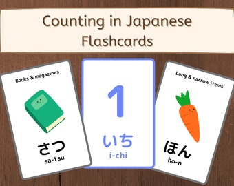 May include: Three flashcards with illustrations and Japanese text. The first card shows a green book with the text "Books & magazines" and the Japanese word "さつ" (sa-tsu). The second card shows the number "1" in blue with the Japanese word "いち" (i-chi). The third card shows a cartoon carrot with the text "Long & narrow items" and the Japanese word "ほん" (ho-n).