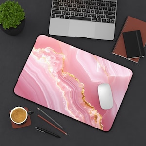 May include: A pink and gold marble-patterned mousepad with a black border. A white computer mouse rests on the pad. A laptop, notebooks, pens, and a cup of coffee are also visible.