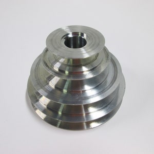 May include: A silver metal pulley with four grooves of different sizes. The pulley has a central hole for mounting.