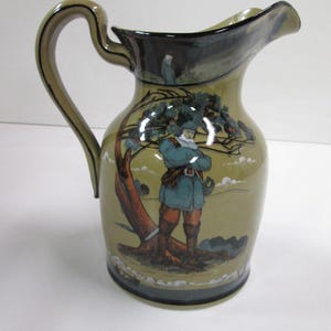 May include: A ceramic pitcher with a handle and spout, featuring a painted scene of a figure in a blue coat and red pants standing near a tree. The pitcher has a green and brown color scheme with a dark rim.