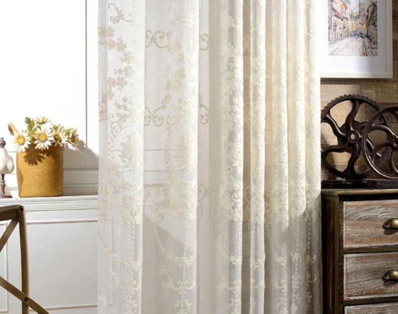 May include: White sheer curtains with an embroidered floral pattern. The curtains are hanging in a window and are letting in natural light.