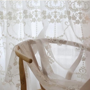May include: White sheer curtains with delicate floral embroidery. The curtains are draped over a wooden chair.