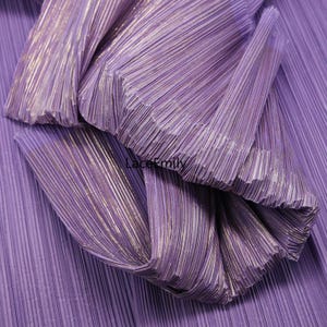 May include: Close-up of pleated fabric in shades of purple with subtle metallic gold accents. The fabric is folded and layered, creating a textured and dimensional effect. The image showcases the intricate details of the pleats and the interplay of light and shadow.