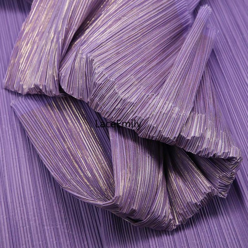 May include: Close-up of pleated fabric in shades of purple with subtle metallic gold accents. The fabric is folded and layered, creating a textured and dimensional effect. The image showcases the intricate details of the pleats and the interplay of light and shadow.