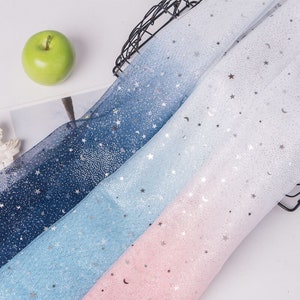 Puede incluir: Tres rollos de tela transparente con un degradado de azul, azul claro y rosa. La tela está decorada con estrellas y lunas plateadas de purpurina.