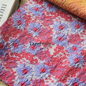 Könnte beinhalten: Nahaufnahme eines weißen Stoffes mit einem rosa-blauen Blumenmuster. Der Stoff hat ein strukturiertes, erhabenes Design, das ihm einen 3D-Effekt verleiht.