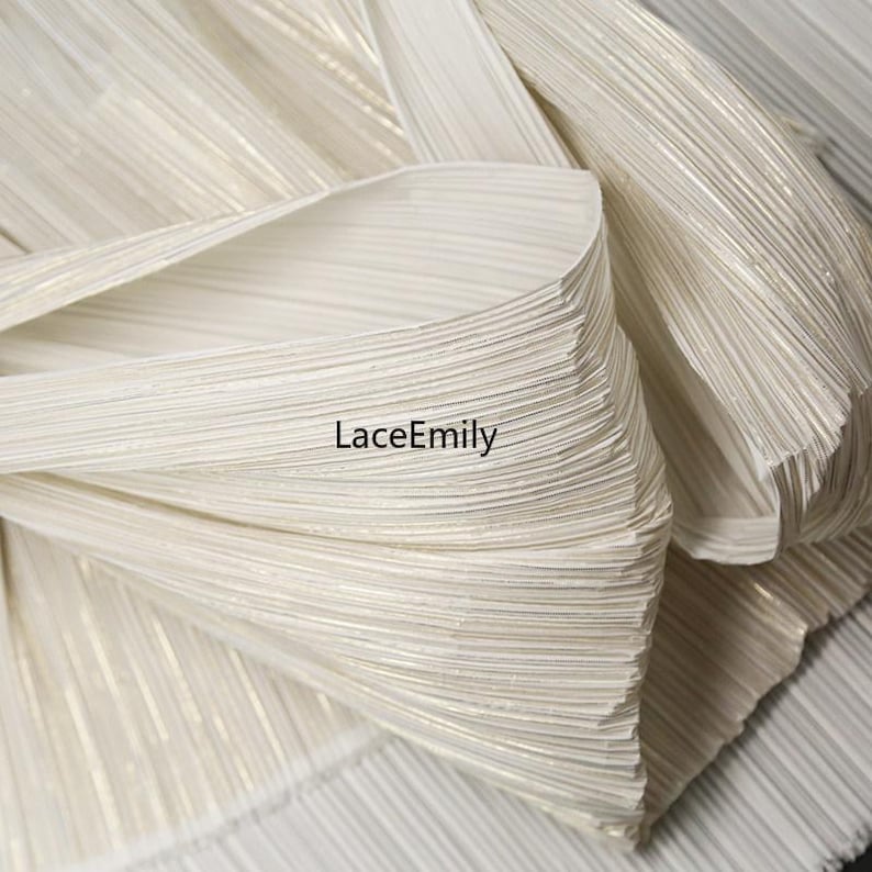 May include: Close-up of a folded, pleated fabric in off-white with subtle gold accents. The fabric has a textured, ribbed appearance, with the brand name "LaceEmily" visible in the image. The material appears to be lightweight and flowing.