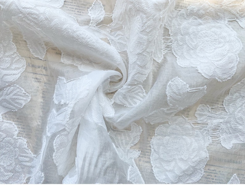 Puede incluir: Tela blanca transparente con un patr&oacute;n floral en relieve. La tela es delicada y tiene una textura suave y fluida.