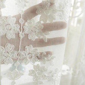 May include: White sheer curtain with delicate floral embroidery. The embroidery is in a white thread and creates a delicate pattern of flowers.