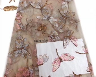 Puede incluir: Tela beige transparente con diseños de libélulas bordadas. Las libélulas están hechas de lentejuelas y cuentas en tonos de rosa, dorado y plateado.