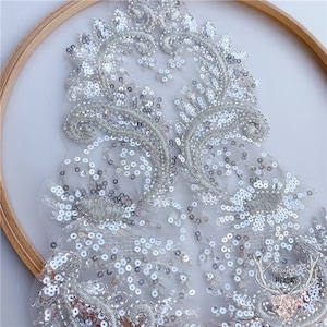 Puede incluir: Aplique de encaje plateado con lentejuelas con un diseño floral. El aplique está sobre un fondo de malla blanca.