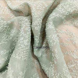 May include: A close-up of a light green sheer fabric with a textured, embossed pattern. The fabric has a delicate, airy feel and is perfect for creating garments or home decor.