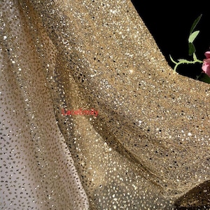 May include: Close-up of shimmering gold fabric with a dense scattering of sequins. The material drapes in soft folds, catching the light and creating a sparkling effect. The fabric has a luxurious appearance, suitable for evening wear or special occasion garments.