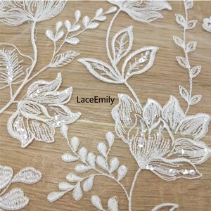 Pu&ograve; includere: Tessuto di pizzo floreale bianco con paillettes. Il tessuto presenta un delicato motivo floreale con piccole foglie e fiori. Il tessuto &egrave; trasparente e ha una texture morbida e fluida.