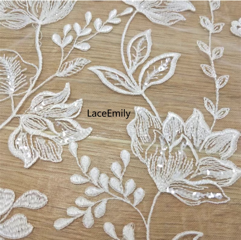 Pu&ograve; includere: Tessuto di pizzo floreale bianco con paillettes. Il tessuto presenta un delicato motivo floreale con piccole foglie e fiori. Il tessuto &egrave; trasparente e ha una texture morbida e fluida.