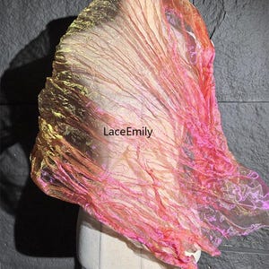 Könnte beinhalten: Ein leichter, halbtransparenter Schal mit knittriger Textur und einem lebendigen Farbverlauf in Gold, Orange und Pink. Der Stoff weist durchgehend einen subtilen irisierenden Glanz auf. Der Text "LaceEmily" ist in Schwarz auf den Schal gedruckt.