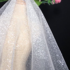 Pode incluir: Tecido de tule branco com glitter prateado. O tecido é transparente e tem uma textura delicada.