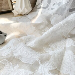 Puede incluir: Tela blanca transparente con un patr&oacute;n floral en relieve. La tela es delicada y tiene una textura suave y fluida.