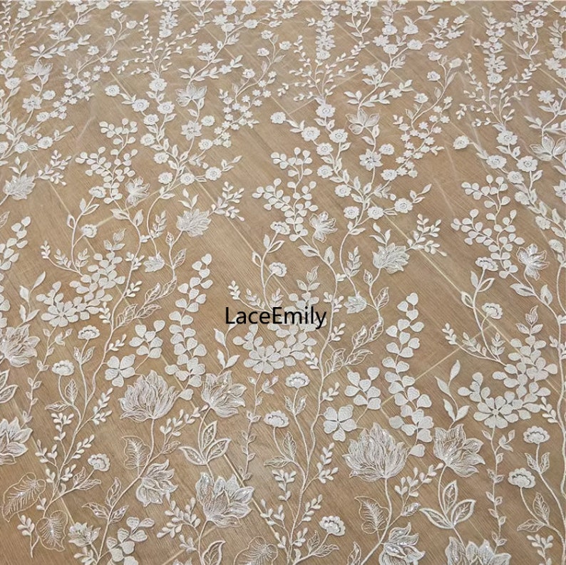Pu&ograve; includere: Tessuto di pizzo floreale bianco con un delicato motivo di fiori e foglie. Il pizzo &egrave; su uno sfondo beige. Il testo "LaceEmily" &egrave; visibile al centro dell'immagine.