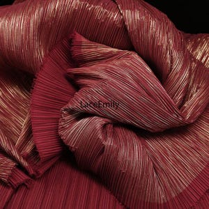 May include: Close-up of a folded, pleated fabric in a deep burgundy color with gold metallic threads running through it. The fabric has a shimmering, textured appearance, with the pleats creating a sense of depth and dimension. The overall effect is luxurious and elegant.