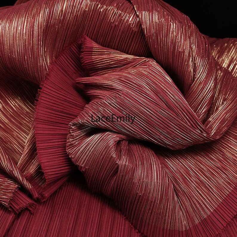 May include: Close-up of a folded, pleated fabric in a deep burgundy color with gold metallic threads running through it. The fabric has a shimmering, textured appearance, with the pleats creating a sense of depth and dimension. The overall effect is luxurious and elegant.
