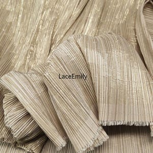 May include: Close-up of gold pleated fabric with a shimmering, textured surface. The fabric is folded, revealing the intricate pleating and a frayed edge. The text "LaceEmily" is visible.