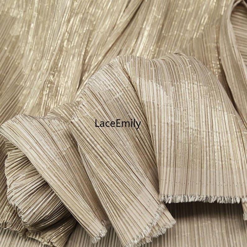 May include: Close-up of gold pleated fabric with a shimmering, textured surface. The fabric is folded, revealing the intricate pleating and a frayed edge. The text "LaceEmily" is visible.