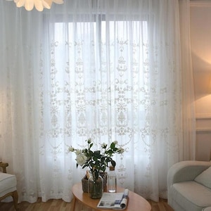 May include: White sheer curtains with an embroidered floral pattern. The curtains are hung in a living room with a couch and a coffee table.