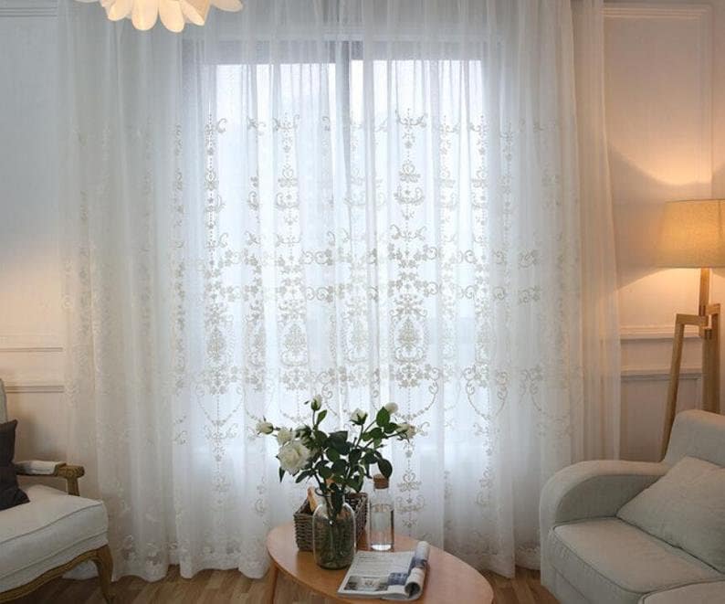 May include: White sheer curtains with an embroidered floral pattern. The curtains are hung in a living room with a couch and a coffee table.