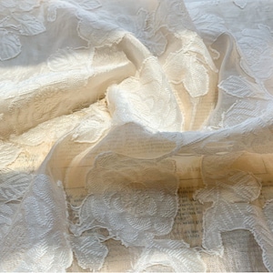 Puede incluir: Tela blanca transparente con un patr&oacute;n floral en relieve. La tela est&aacute; drapeada y doblada, creando una textura suave y delicada.