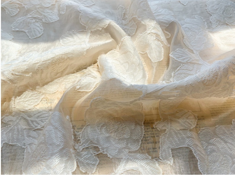 Puede incluir: Tela blanca transparente con un patr&oacute;n floral en relieve. La tela est&aacute; drapeada y doblada, creando una textura suave y delicada.