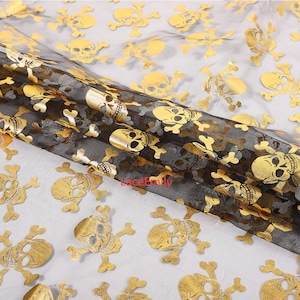 May include: Black and gold skull and crossbones fabric. The fabric is sheer, with a black base and gold foil skull and crossbones pattern. The skulls are detailed and evenly spaced across the fabric.