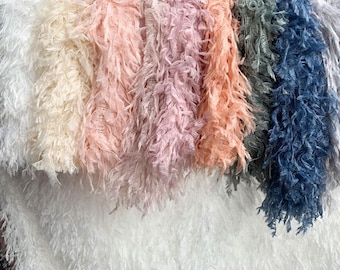 May include: A collection of fringed fabric strips arranged in a vertical row, featuring a gradient of colors including white, cream, pale pink, dusty rose, peach, sage green, navy blue, and grey, each with a textured, shaggy, and fibrous appearance.