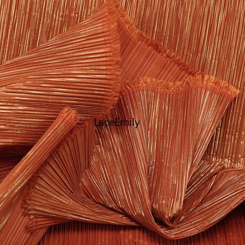 May include: Close-up of a folded, pleated fabric in a warm, copper-orange hue. The fabric has a vertical ribbed texture, creating a shimmering effect. The image shows the fabric's folds and edges, highlighting its texture and sheen. The text "LaceEmily" is visible.