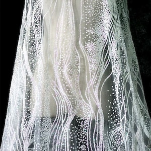 Pode incluir: Tecido branco com glitter iridescente com um padrão ondulado. O tecido é transparente e tem uma textura delicada.