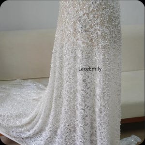May include: Close-up of a white, sequined fabric, likely for bridal or formal wear. The material has a flowing drape and a shimmering texture. The text "LaceEmily" is visible on the fabric.