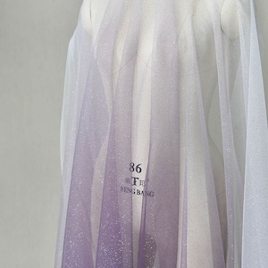 May include: A sheer, iridescent fabric with a gradient of purple to white. The fabric is covered in tiny silver glitter. The fabric is labeled "86° Feng Bang".