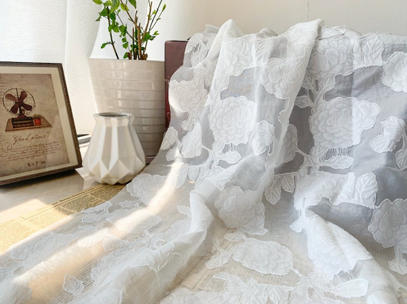 Puede incluir: Tela blanca transparente con un patr&oacute;n floral en relieve. La tela est&aacute; drapeada sobre una superficie y tiene un aspecto delicado y rom&aacute;ntico.