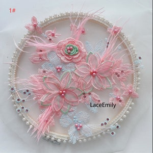 May include: A pink and white floral embroidery applique with pearl beads and feathers. The applique is circular and has a delicate lace design. The text "LaceEmily" is visible in the center of the applique.