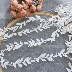 May include: White lace trim featuring a delicate leaf design. The trim is displayed wrapped around a wooden embroidery hoop.