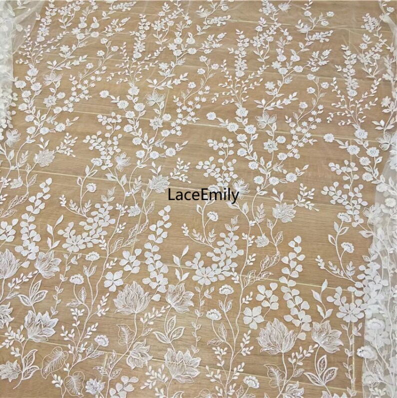 Pu&ograve; includere: Tessuto di pizzo floreale bianco con un delicato motivo di fiori e foglie. Il tessuto &egrave; trasparente e ha una texture morbida e fluida. Il testo "LaceEmily" &egrave; visibile al centro dell'immagine.